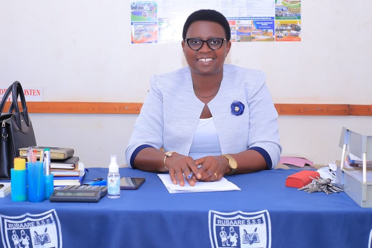 Headteacher Ms. Mpeirwe Monic Atukunda wearing glasses and seated at her desk with Rubaare Secondary School branding visible.